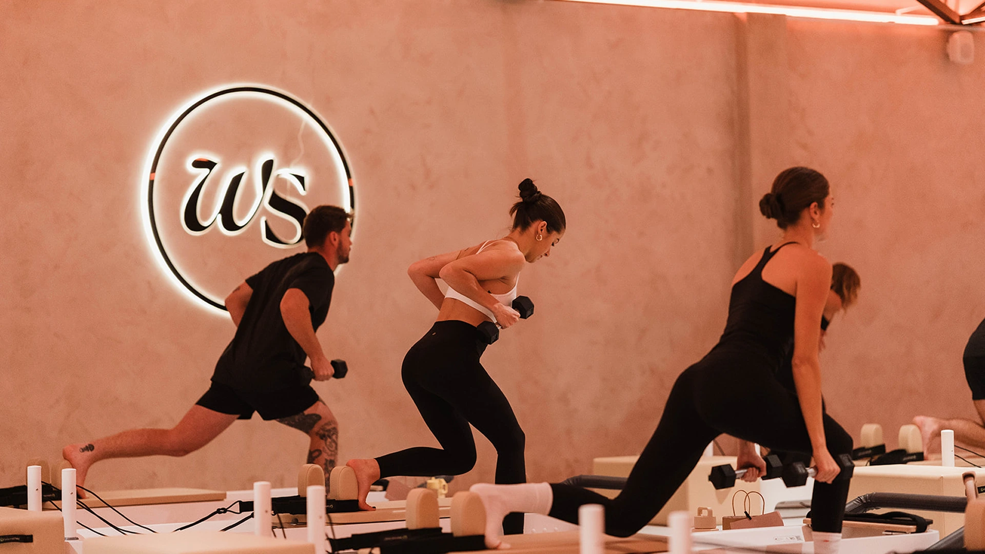 People in activewear performing low lunges while holding dumbbells during a fitness class in a warm-lit studio with a glowing wall logo.