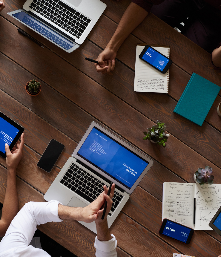 Top-down view of team members working around a wooden conference table with laptops, tablets, smartphones, and notebooks