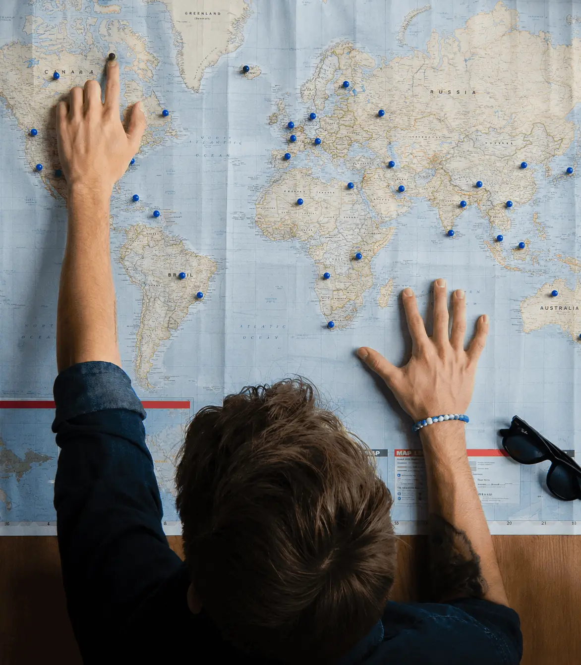 Top-down view of a man reaching over a large world map marked with blue push pins, pointing his finger toward North America.