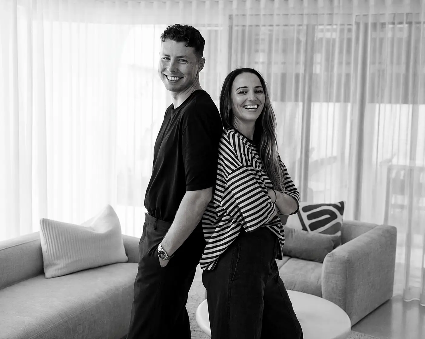 Black and white portrait of a smiling man and woman standing back-to-back in a modern living space with curtains and sofas in the background.