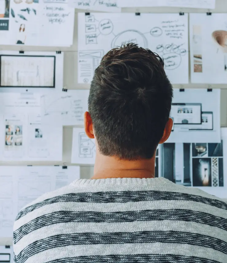 Back view of a man in a striped sweater standing in front of a white wall filled with pinned wireframes, sketches, and planning notes.